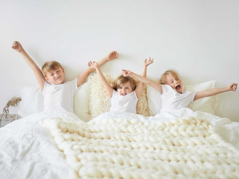 Bedding, sleep, rest and relaxation concept. Indoor shot of three children feeling sleepy while waking up early in the morning before school. Two brothers and sister yawning and stretching in bed