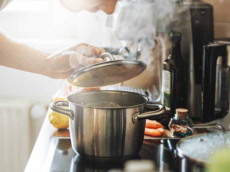 Young man cooking fresh food at home and opening lid of steaming pot.